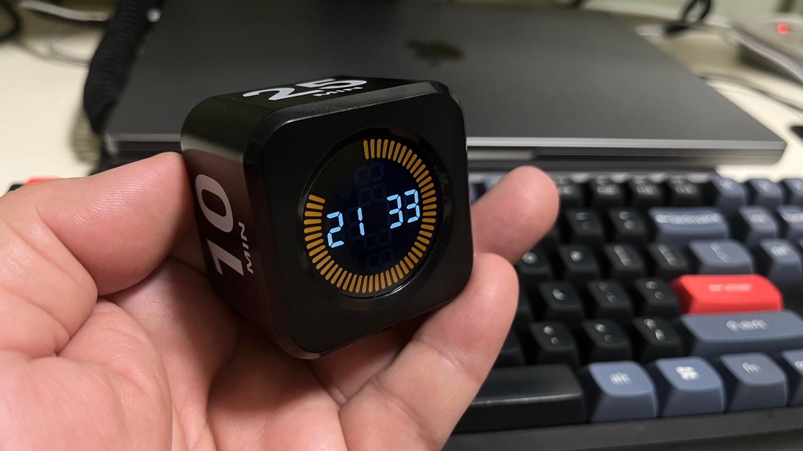A black cube-shaped Pomodoro timer showing 21:33 on its display, resting in a hand next to a mechanical keyboard and a laptop.